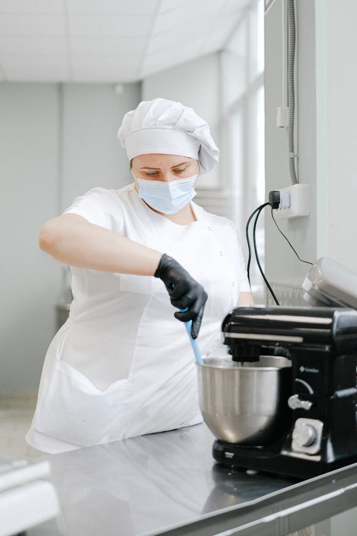 Home A chef wearing protective gear mixes ingredients using a stand mixer in a commercial kitchen.